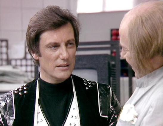 Claude 3.7 describes the image as: "The image shows two people in conversation in what appears to be a futuristic or science fiction setting. On the left is someone wearing a distinctive black outfit with a metallic or studded vest/jacket with silver accents. They have dark hair and are speaking to another person on the right who is wearing lighter-colored clothing, possibly white or cream.

The scene appears to be from a science fiction television production from the late 1970s or early 1980s …