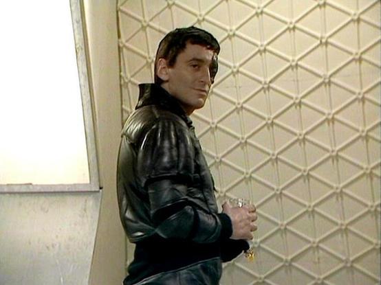 Claude 3.7 describes the image as: "The image shows a person in a black leather jacket standing in what appears to be an interior setting with a distinctive textured wall featuring a diamond pattern in the background. The individual is turned slightly, looking over their shoulder, and appears to be holding something small in their hands. The setting has a somewhat institutional or utilitarian appearance with simple walls and minimal decoration, which is characteristic of the retro sci-fi aesthe…