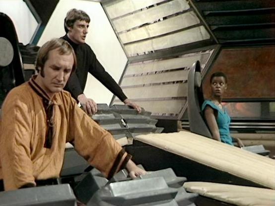 Claude Sonnet 4.0 describes the image as: "This scene appears to be set aboard the Liberator, the advanced alien spacecraft that serves as the main setting for much of the series. The interior shows the characteristic white and geometric design of the ship's living areas or flight deck. Three crew members are present in what appears to be a tense or serious moment. One character is seated wearing earth-toned clothing, while another stands behind in dark attire. A third crew member can be seen i…