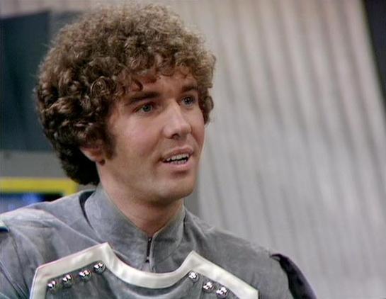 Claude 3.7 describes the image as: "This image appears to be from a science fiction television series, showing a person with curly brown hair wearing what looks like a futuristic silver or gray costume with armored elements. The character is in what appears to be a spacecraft or technical setting, with light-colored panels visible in the background. The costume has a distinctive metallic collar and chest plate with some small decorative elements or controls. The styling and production design su…
