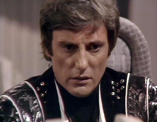 Claude 3.7 describes the image as: "The image shows a person in what appears to be a science fiction television production from the late 1970s or early 1980s, based on the filming style and costume design. 

The individual is wearing a distinctive black leather outfit with metallic studs or embellishments, which is characteristic of the futuristic, somewhat dystopian aesthetic common in British sci-fi of that era. 

The costume has a militaristic or authoritarian quality, with structured should…