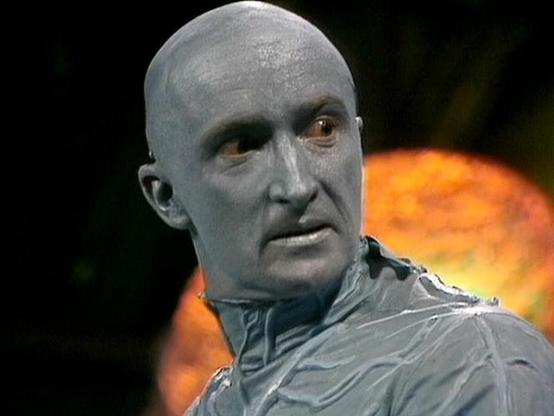 Claude Sonnet 4.0 describes the image as: "This image shows a character with distinctive pale, greyish makeup and golden-colored eyes, wearing what appears to be a metallic or silver-colored garment. The character has a bald head and an otherworldly appearance that suggests they are an alien or artificially enhanced being. The background features warm orange and reddish lighting, creating a dramatic sci-fi atmosphere typical of the series. The makeup and costume design emphasize the character's…