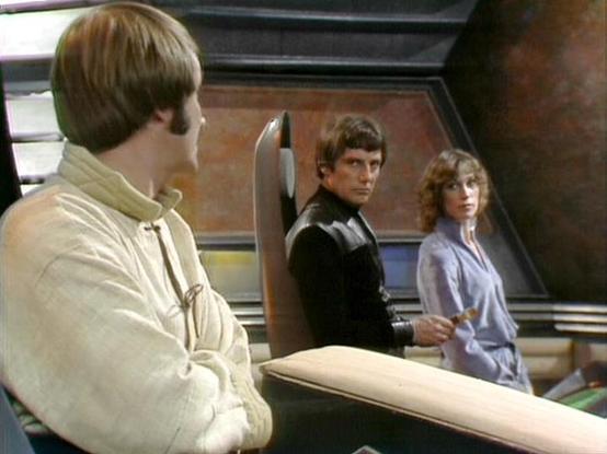 Claude 3.7 describes the image as: "This image appears to be from a science fiction television show from the late 1970s or early 1980s based on the production style and costume design. The scene is set in what looks like a spacecraft or control room with metallic and brown paneled walls.
The image shows three people in a tense moment. On the left is someone in a light-colored, high-collared outfit. In the center and right of the frame are two others - one wearing dark clothing and another in a…