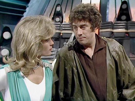 Claude Sonnet 4.0 describes the image as: "I can see this is a scene from a science fiction television series featuring two characters in what appears to be a futuristic spacecraft setting. The background shows typical sci-fi television production design with metallic surfaces and circular lighting elements. One character is wearing a mint green and white outfit with a chain necklace and has blonde hair, while the other wears earth-toned clothing including what appears to be a leather jacket ov…