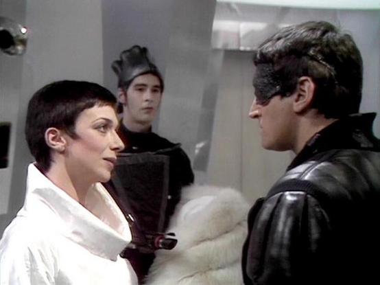 Claude 3.7 describes the image as: "# Scene Description

This image appears to be from a science fiction television production, set in what looks like a minimalist, futuristic interior space with plain gray walls and metallic fixtures. The setting has a sterile, institutional quality typical of 1970s-80s sci-fi design.

In the foreground, a woman wearing a white garment with dark trim is engaged in conversation with a man in a black leather jacket. Between them appears to be some kind of white …