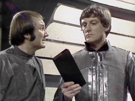 Claude Sonnet 4.0 describes the image as: "This image appears to be from the British science fiction television series "Blake's 7," which aired from 1978 to 1981. The scene shows two characters in what looks like a spacecraft interior, with distinctive white paneled walls featuring horizontal stripes in the background.

On the left, Michael Keating portrays Vila Restal, a character known as the ship's thief and lock-breaker. He's wearing a grey tunic-style costume typical of the show's utilitar…