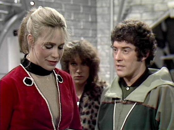 Claude Sonnet 4.0 describes the image as: "This image appears to be from a classic British science fiction television series, likely from the late 1970s or early 1980s based on the production quality and costume design. The scene shows three characters in what appears to be a spaceship or futuristic facility interior, with industrial gray walls visible in the background.

The woman on the left wears a distinctive red velvet tunic with white cord trim and a black turtleneck underneath, styled wi…