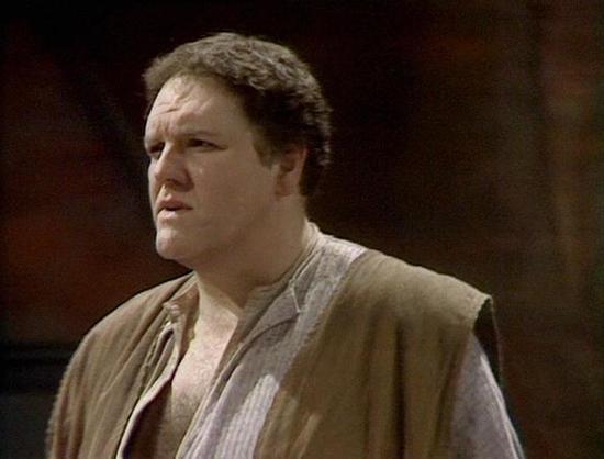 Claude Sonnet 4.0 describes the image as: "This image shows a scene from what appears to be a television production, likely from the late 1970s or early 1980s based on the video quality and styling. The setting appears to be indoors with warm, dim lighting creating a dramatic atmosphere. The person in the scene is wearing period-appropriate clothing consisting of earth-toned, loose-fitting garments that suggest either a historical drama or science fiction setting. The costume includes what appe…