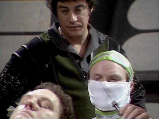 Claude 3.7 describes the image as: "# Scene Description

This image appears to be from a science fiction television production, showing a medical or clinical setting. The scene captures three individuals in what looks like a spacecraft or futuristic facility interior, characterized by the metallic walls visible in the background.

In the foreground, a person wears a distinctive lime green headband and white medical mask, suggesting a medical or emergency scenario. Behind them, another figure in…
