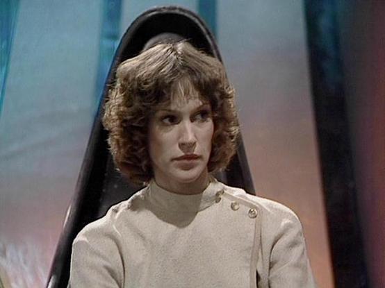 Claude Sonnet 4.0 describes the image as: "This image shows a scene from the British science fiction television series "Blake's 7," which aired from 1978 to 1981. The photograph captures Jan Chappell in character as Cally, a telepathic alien from the planet Auron. She wears a cream-colored, high-necked uniform with distinctive shoulder buttons, characteristic of the show's utilitarian costume design. Her shoulder-length curly hair and direct gaze convey the character's serious, contemplative na…