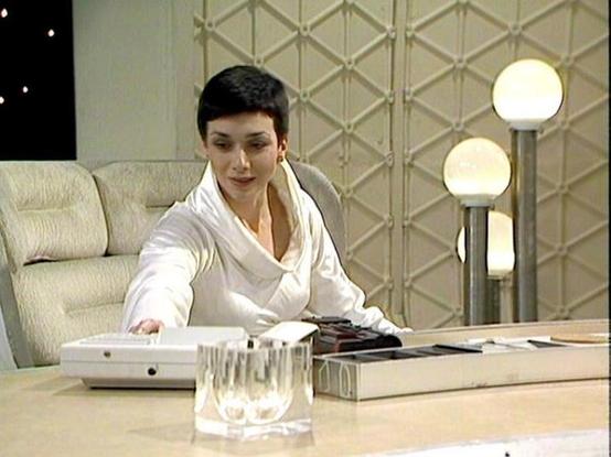 Claude 3.7 describes the image as: "The image shows a person in a white outfit seated at a desk or table in what appears to be a stylized, retro-futuristic set with a distinctive geometric wall pattern in the background. The setting features round globe lights mounted on a stand and a light-colored couch or seating area visible to the left.
The person has short dark hair and is reaching toward what looks like office equipment on the desk, possibly a communication device. There's also what appe…