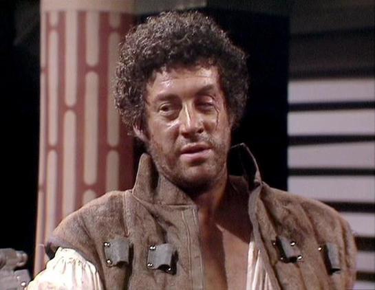 Claude Sonnet 4.0 describes the image as: "This image shows a dramatic scene from what appears to be a science fiction television production, likely from the 1970s based on the visual style and production quality. The actor Gareth Thomas is portrayed in period costume consisting of a rustic brown leather or suede vest over a billowing white shirt, giving him the appearance of a historical or adventurous character. His dark, curly hair frames his face as he gazes intensely off-camera, suggesting…