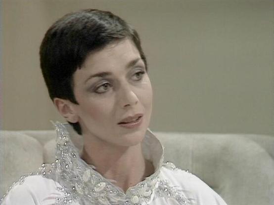 Claude 3.7 describes the image as: "The image shows a person with short dark hair in what appears to be a scene from a television production. They are wearing an elegant white outfit with a decorative crystal or beaded collar that has a distinct vintage sci-fi or fantasy aesthetic. The individual has a contemplative or mid-conversation expression and is seated against what looks like a light-colored sofa or seating area.

The setting appears to be from a classic television production, likely fr…