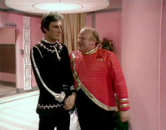 Claude Sonnet 4.0 describes the image as: "This image shows two actors in elaborate costume standing in a distinctly retro-futuristic set with pink walls and modern lighting. The setting appears to be from a British science fiction production, likely from the 1970s or early 1980s, characterized by its bright pastel color scheme and geometric design elements.

Paul Darrow, on the left, wears a striking black uniform with silver trim and ornate detailing, including decorative elements along the s…