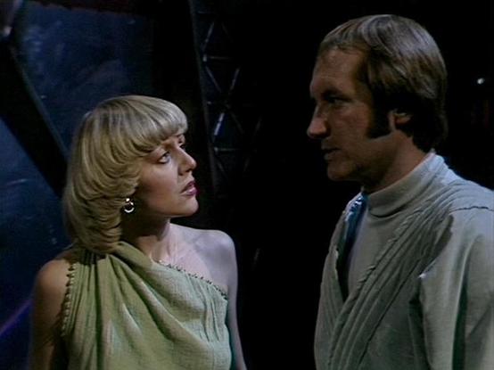 Claude Sonnet 4.0 describes the image as: "This image appears to be from the British science fiction television series "Blake's 7," which aired from 1978 to 1981. The scene shows two characters in what appears to be a spaceship or futuristic setting, with dark metallic walls visible in the background. Both individuals are wearing flowing, draped clothing in muted earth tones that's characteristic of the show's costume design. The woman has a blonde, layered hairstyle typical of late 1970s fashi…