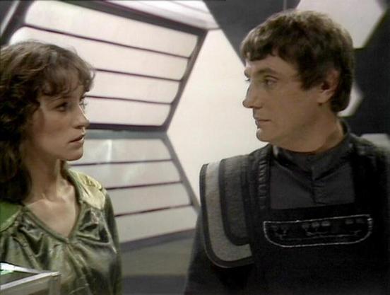 Claude 3.7 describes the image as: "This image appears to be from the classic British science fiction television series "Blake's 7." The scene shows two people in a futuristic spacecraft setting, with distinctive angular wall panels visible in the background that were characteristic of the show's set design.

The person on the left is wearing a greenish metallic top, while the person on the right is dressed in a dark outfit with a structured vest or armor piece that has padded shoulders and a p…
