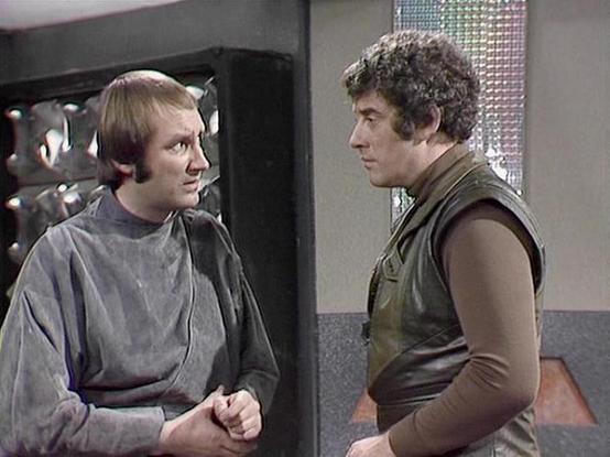 Claude 3.7 describes the image as: "This image appears to be from a science fiction television series, likely from the late 1970s or early 1980s based on the production style and costumes. The scene shows two individuals in conversation within what looks like a spacecraft or futuristic facility with metallic walls and reflective surfaces visible in the background.

The person on the left is wearing a gray uniform-like outfit, while the person on the right is dressed in a brown/olive sleeveless …