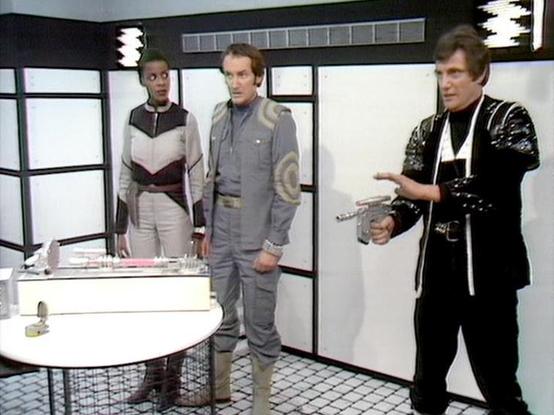 Claude 3.7 describes the image as: "This image appears to be from the classic British science fiction series "Blake's 7." The scene takes place in what looks like a futuristic spacecraft interior with white walls, translucent panels, and a minimalist design aesthetic typical of sci-fi productions from that era.

Three people are shown in distinctive costume designs that reflect the show's characteristic styling. On the left, a person wears a two-tone outfit with a chevron pattern. In the center…