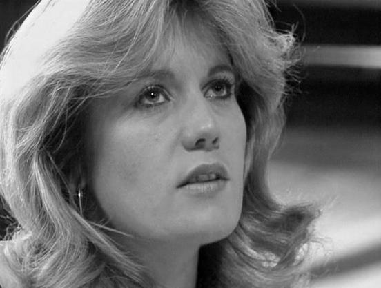 Claude 3.7 describes the image as: "This is a black and white close-up portrait showing a person with feathered, shoulder-length hair styled in a 1970s fashion. The image captures an intense, thoughtful expression with the subject gazing slightly upward. The lighting creates a dramatic contrast that emphasizes the facial features and the texture of the hair. The background is blurred but appears to be a simple, dark setting typical of film or television production from that era.
The monochrome…