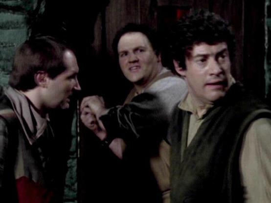 Claude 3.7 describes the image as: "# Scene Description

This image appears to be from a television production, showing three men in what looks like an indoor setting with dark, industrial-style surroundings. The lighting is dim and atmospheric, suggesting either a spacecraft interior or underground facility.

The men are dressed in casual clothing - one wears a red and beige/tan colored outfit on the left, while the others wear earth-toned garments including browns and grays. Their positioning…