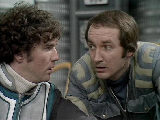 Claude Sonnet 4.0 describes the image as: "This image shows two actors in what appears to be a science fiction television production, likely from the late 1970s or early 1980s based on the styling and image quality. Both men are wearing futuristic costumes - one in a white and teal outfit with distinctive collar details, and the other in earth-toned brown and green garments. The setting appears to be a spaceship or futuristic facility interior, with metallic surfaces and technological equipment…