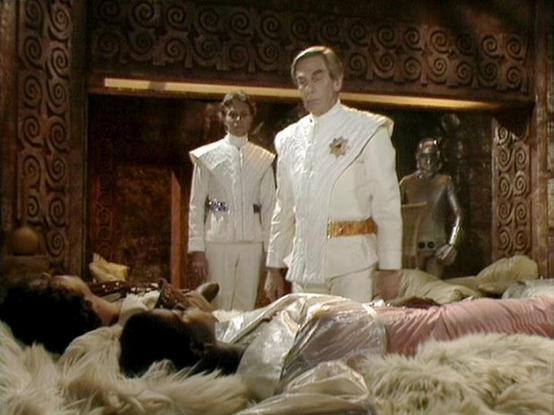 Claude Sonnet 4.0 describes the image as: "This scene appears to be from a science fiction television production, likely from the late 1970s or early 1980s based on the visual quality and costume design. The setting is an ornate, luxurious chamber with decorative spiral patterns on dark walls and ambient lighting creating an atmospheric mood.

Two figures stand prominently in white ceremonial or military-style uniforms with gold accents and insignia, positioned authoritatively in the center of …