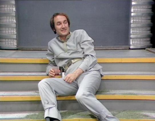 Claude 3.7 describes the image as: "# Image Description

This image shows a man in a light gray/beige tracksuit sitting casually on stadium bleachers with yellow and gray striping. He's positioned in a relaxed pose with one leg extended, holding what appears to be a beverage container in his hand. The setting appears to be an indoor or covered stadium area, with metal railings visible in the background against a blue-gray wall.

The man has shoulder-length brown hair styled in a way characteris…