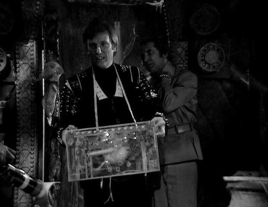 Claude 3.7 describes the image as: "# Black and White Scene Analysis

This black and white image appears to be from a science fiction production, showing two figures in an ornate, elaborate setting. The man on the left wears a distinctive black outfit adorned with studded details and decorative elements, while another figure stands to his right in a more utilitarian brown/gray uniform. The background features intricate decorative patterns and geometric designs that suggest an alien or futuristi…