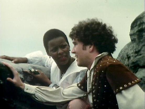 Claude 3.7 describes the image as: "# Image Description

This appears to be a scene from a film or television production, likely from the 1980s based on the video quality and styling. Two actors are shown in an outdoor setting against a rocky backdrop. One performer wears a white shirt and appears engaged in conversation with the other, who is dressed in a brown leather vest decorated with metal studs or grommets, suggesting a period or fantasy costume.

The scene captures an intimate moment of…