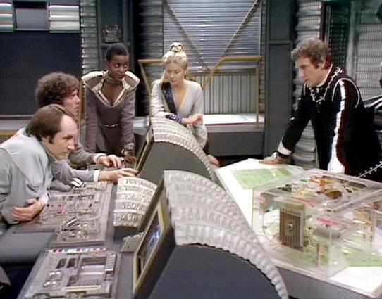 Claude Sonnet 4.0 describes the image as: "This image shows a scene from the British science fiction series "Blake's 7," featuring five actors gathered around a futuristic control console. The setting appears to be the flight deck of the spacecraft Liberator, with its distinctive curved control panels and technical equipment visible throughout.

The actors are engaged in what appears to be a tactical discussion, gathered around illuminated control surfaces displaying maps or strategic informati…