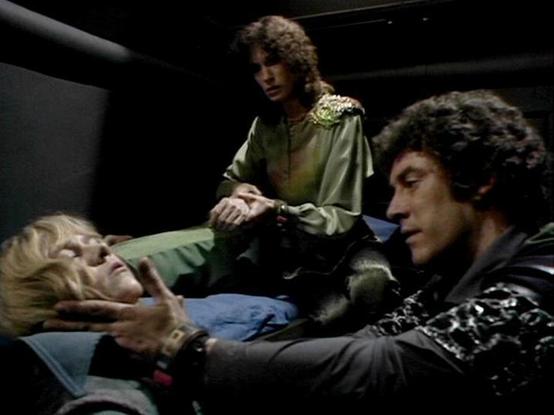 Claude Sonnet 4.0 describes the image as: "This appears to be a dramatic scene from the British science fiction television series "Blake's 7." The image shows three actors in what looks like a tense medical or crisis situation aboard a spacecraft. A blonde woman lies on what appears to be a medical bed or examination table, seemingly unconscious or in distress. Standing over her in a green satin-like outfit with ornate shoulder detailing is one actor, while another wearing a patterned vest lean…