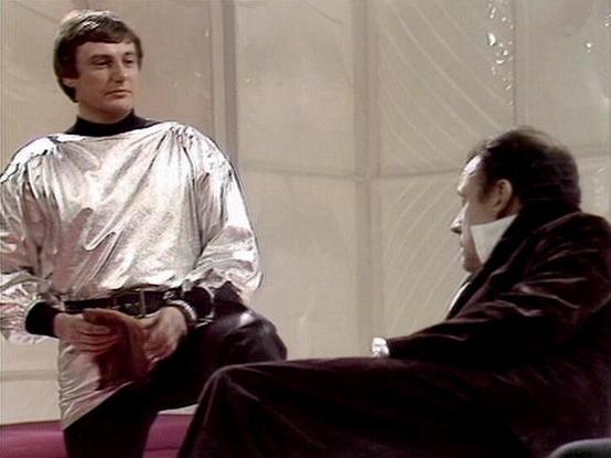 Claude Sonnet 4.0 describes the image as: "This scene appears to be from a science fiction television production, likely from the 1970s or early 1980s based on the visual quality and costume design. The setting features a sleek, minimalist interior with pale walls and geometric patterns, characteristic of futuristic set design from that era.

In the foreground, one actor wears a distinctive metallic silver tunic with voluminous sleeves over black clothing, creating a striking space-age appearan…