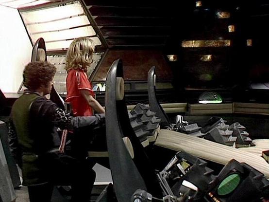 Claude 3.7 describes the image as: "This image shows a scene from what appears to be the classic British science fiction series "Blake's 7." The setting is the interior of a spacecraft, likely the flight deck of the Liberator, with its distinctive control panels and futuristic design elements. 

The scene depicts two people in the ship's control room - one wearing dark clothing with a green garment, and another in a red outfit. They appear to be operating or examining the ship's controls. The s…
