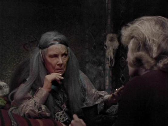 Claude Sonnet 4.0 describes the image as: "This appears to be a scene from a period drama or horror film, set in what looks like a dimly lit, rustic interior. The atmosphere is moody and theatrical, with dramatic lighting creating shadows throughout the scene. Two figures are visible - one appears to be an older woman with long, grey hair wearing period costume with ornate details, seated and appearing to be in conversation or consultation. The other figure is partially visible in the foregroun…