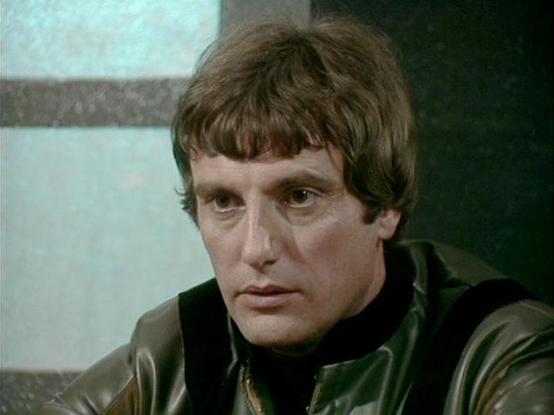 Claude 3.7 describes the image as: "This image shows a man with dark brown hair styled in a 1970s fashion, featuring bangs and shoulder-length layers. He's wearing a dark leather or vinyl jacket over what appears to be a black shirt or turtleneck. The setting appears to be indoors, with a muted, institutional-looking background featuring green and pale blue tones typical of television studio sets from that era.

The lighting and color palette suggest this is from a science fiction television pr…