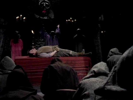 Claude 3.7 describes the image as: "# Theatrical Scene Analysis

This appears to be a dramatic stage production set in a gothic or medieval environment. The scene depicts a funeral or death ritual, with a figure lying horizontally on a red fabric-draped platform or altar. The setting is notably dark, with minimal lighting that creates an eerie atmosphere.

The staging includes numerous figures dressed in dark, period-appropriate clothing standing solemnly in the background. Some figures hold ca…