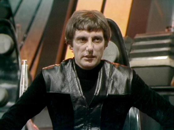 Claude Sonnet 4.0 describes the image as: "This image shows a scene from the classic British science fiction television series "Blake's 7" from the late 1970s/early 1980s. The setting appears to be the flight deck or control room of the alien spacecraft Liberator, recognizable by its distinctive curved architectural elements and control panels with a bronze/orange color scheme. The character is wearing the iconic black leather outfit typical of the show's rebel crew members. The scene captures …