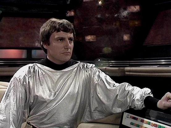 Claude 3.7 describes the image as: "# Scene Description

This image shows a figure in a silvery, flowing tunic-style garment seated at what appears to be a control console or command station. The setting features a futuristic interior with dark, metallic walls and atmospheric lighting in warm tones. Behind the figure is a sleek panel with horizontal lines characteristic of science fiction production design from the 1970s-1980s era.

The character displayed a composed, serious demeanor while sea…