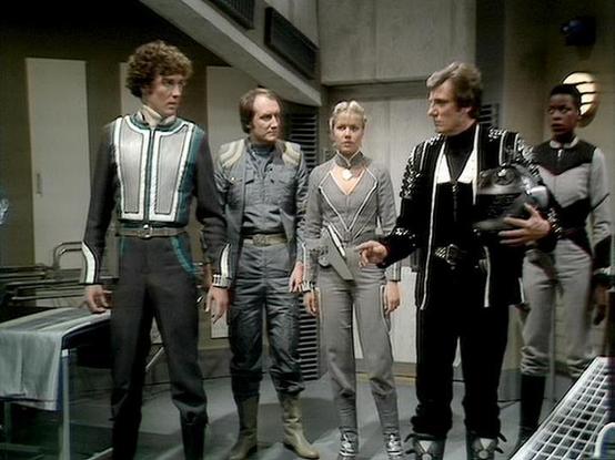 Claude 3.7 describes the image as: "This image appears to be from the classic British science fiction television series "Blake's 7" from the late 1970s/early 1980s. The scene shows a group of people in futuristic costumes standing in what looks like a spacecraft or space station interior with gray walls and utilitarian design.

The costumes feature distinctive styling typical of sci-fi productions of that era - including a character in black leather with silver accents holding what appears to b…