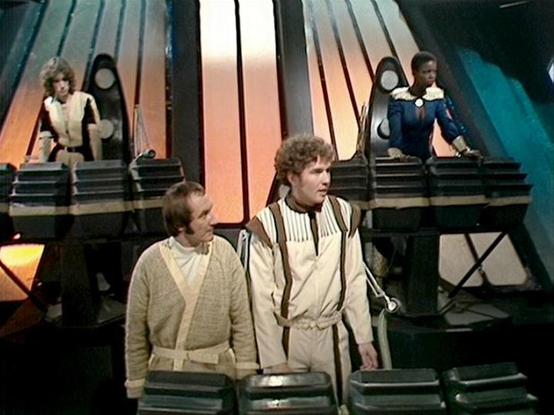 Claude 3.7 describes the image as: "This image shows a scene from the classic British science fiction TV series "Blake's 7," set on what appears to be a spacecraft bridge or control room. The setting features a futuristic design with angular metallic panels and control consoles typical of the show's distinctive visual style from the late 1970s/early 1980s.

The crew members are positioned at different levels of the set - two at control stations in the background on elevated platforms, while two…
