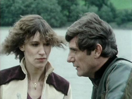 Claude 3.7 describes the image as: "This image appears to be a still from a vintage television production, likely from the late 1970s or early 1980s based on the film quality and styling. The scene shows two people in conversation outdoors near what seems to be a body of water, with greenery in the background. The setting has a somewhat muted, cool color palette typical of productions from that era.

The person on the left is wearing a light-colored jacket with darker accents, while the person …