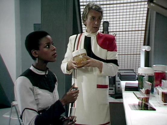 Claude Sonnet 4.0 describes the image as: "This appears to be a scene from a science fiction television production, likely from the 1980s based on the aesthetic and production values. The setting is a futuristic medical facility or laboratory, characterized by clean white surfaces, advanced-looking equipment, and sterile lighting typical of sci-fi productions from that era.

The scene shows two characters in what appears to be a medical or scientific consultation. One character is seated, weari…