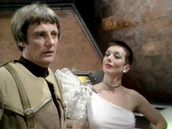 Claude 3.7 describes the image as: "This image appears to be from the classic British science fiction television series "Blake's 7." The scene shows two performers in costume on what looks like a futuristic set with textured walls typical of the show's aesthetic.

On the left is a person wearing a beige/brown outfit with ribbed details and suspenders, with short dark hair. On the right is a person in an elegant white one-shoulder dress with a distinctive silver collar or necklace and very short…
