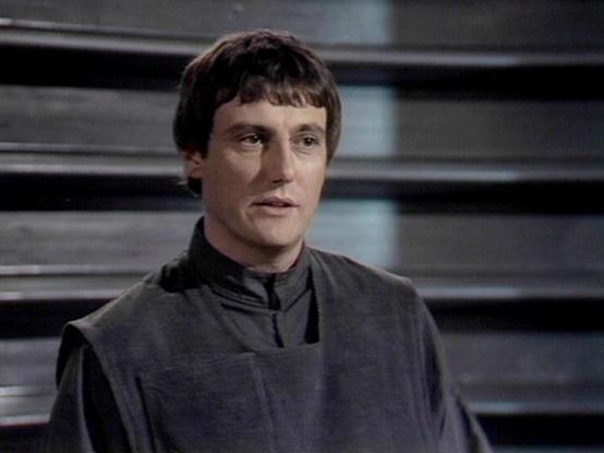 Claude Sonnet 4.0 describes the image as: "This image shows a scene from what appears to be a British science fiction television production, likely from the late 1970s or early 1980s based on the visual quality and aesthetic. The setting features a stark, industrial backdrop with horizontal metal or plastic paneling creating a futuristic corridor or spaceship interior.

Paul Darrow appears in character wearing a distinctive grey-black tunic with a high collar characteristic of utilitarian sci-f…