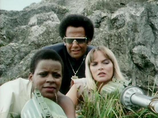 Claude 3.7 describes the image as: "# Image Description

This photograph captures three people posing together outdoors against a rocky, textured cliff face backdrop. The setting appears to be a natural, rugged landscape with grey stone formations visible behind them.

The composition features three individuals arranged casually for the camera. On the left is a person wearing a light-colored shirt. In the center stands a man wearing dark clothing, a chain necklace, and distinctive oversized sun…