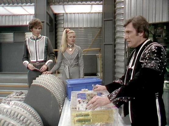 Claude 3.7 describes the image as: "This image appears to be from the classic British science fiction series "Blake's 7." The scene takes place in what looks like a spacecraft or space station interior with metallic walls and industrial styling typical of the show's production design.

Three people are gathered around what appears to be a control console or workstation. On the right, a person wearing a distinctive black outfit with decorative elements stands at the console, appearing to operate…