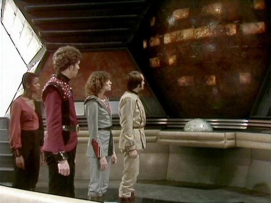 Claude 3.7 describes the image as: "# Blake's 7 - Observation Dome Scene

This image depicts a scene from the science fiction series Blake's 7, showing four characters standing in what appears to be a spacecraft's observation dome or bridge area. The setting features futuristic architecture with metallic walls and a large viewing screen displaying what looks like a planetary body or celestial object.

The characters are dressed in the distinctive costume design of the 1970s-80s sci-fi series, w…