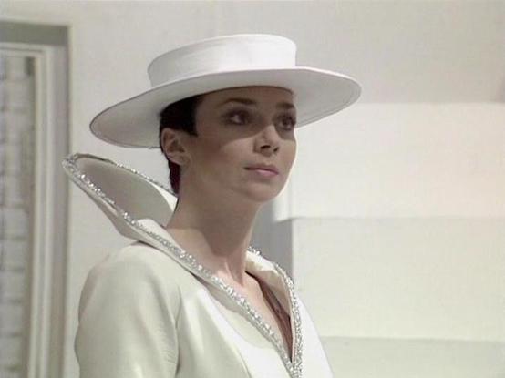 Claude 3.7 describes the image as: "This image shows a scene from what appears to be a science fiction television production, likely from the British series "Blake's 7." The actor Jacqueline Pearce is dressed in an elaborate white costume featuring a distinctive wide-brimmed white hat and a high, sculptural collar embellished with what appears to be silver beading or sequins. The costume has a regal, futuristic quality that suggests a character of authority or high status.

The setting appears …