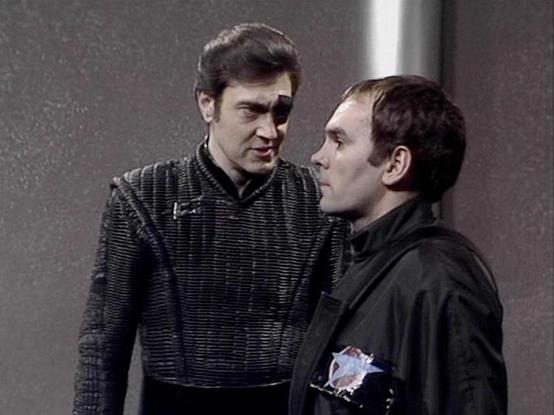 Claude Haiku 4.5 describes the image as: "I can see two men in what appears to be an interior setting with plain walls and minimal background detail. The man on the left wears a dark textured sweater or shirt, while the man on the right wears a black jacket with what appears to be a small insignia or patch on the chest. They are positioned facing each other in what seems to be a conversational or confrontational moment. The lighting and image quality suggest this is from a television production…