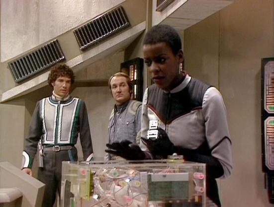 Claude Sonnet 3.7 describes the image as: "The image shows a scene from the British science fiction television series "Blake's 7." It depicts three crew members in a futuristic corridor setting aboard their spacecraft. They are wearing distinctive costume designs typical of the show's aesthetic, with structured uniforms featuring color blocking and geometric patterns in grays, whites, and accent colors.

In the foreground, a crew member is holding what appears to be a transparent container or d…